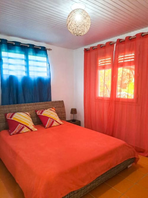 Chambre lumineuse à la Villa Tourmaline avec lit rouge, coussins rayés, tête de lit en rotin et rideaux rouges au Lorrain