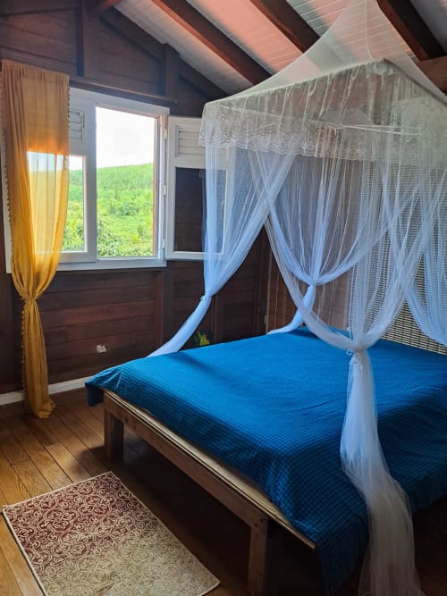 Chambre de la Villa Tourmaline au Lorrain avec lit à baldaquin bleu, voilages blancs et vue sur la rivière verdoyante