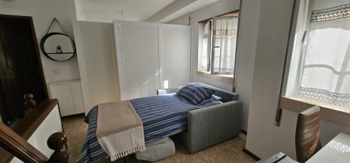 Quarto luminoso em Sunao Nazaré com cama de listras azuis, cabeceira estofada cinzenta, espelho redondo e janelas com luz natural