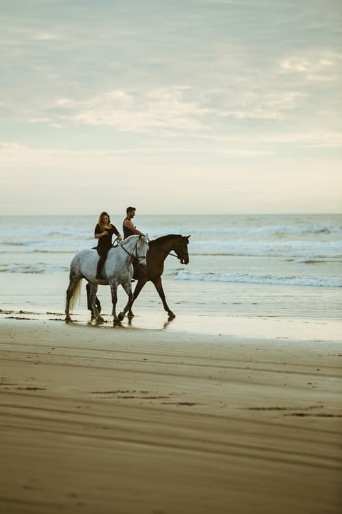 Dos jinetes a caballo trotando en la playa de Chipiona con olas y cielo al fondo
