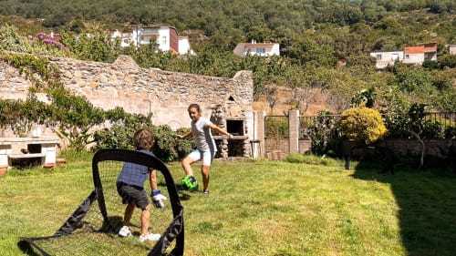 Niños jugando al fútbol en el jardín verde de Casa Rural Villa del Jerte, El Torno Cáceres, con edificios rústicos cercanos