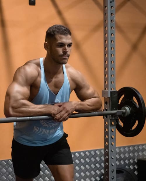 Homme musculaire en débardeur bleu tenant une barre de musculation dans une salle de sport avec fond orange