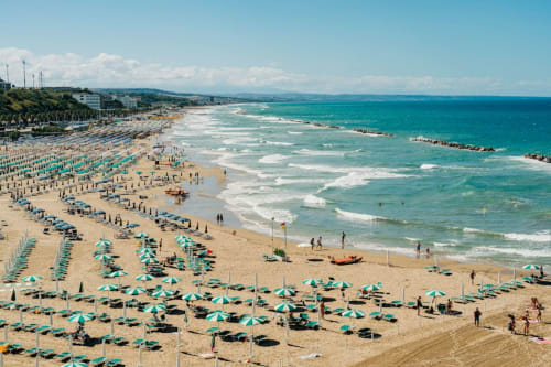 Ombrelloni turchesi punteggiato la spiaggia sabbiosa di San Giacomo degli Schiavoni con acque cristalline del Mediterraneo e edifici costieri