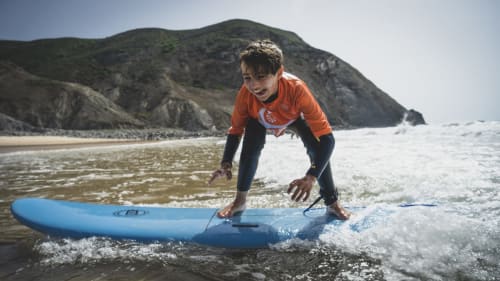 Young surfer in orange wetsuit riding wave at Bordeira beach with dramatic cliffs backdrop