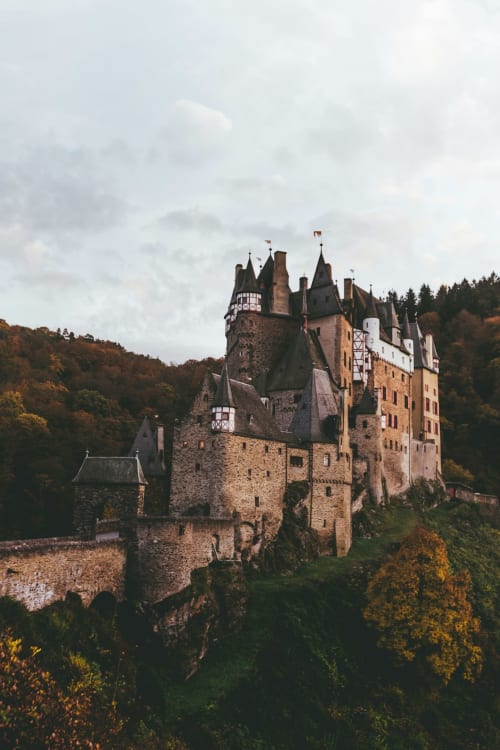 Medieval castle nestled in forested valley with stone towers and turrets under cloudy sky