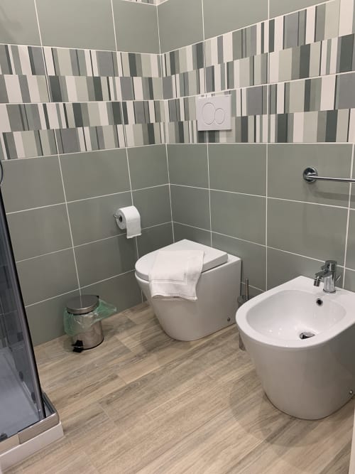 Modern bathroom at Maryam 23 B&B in Siracusa with gray tiled walls, white toilet, bidet, sink, and wood-look flooring