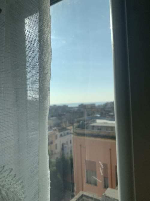 City view from Maryam 23 B&B window in Siracusa, with terracotta rooftops and clear sky visible through sheer curtains