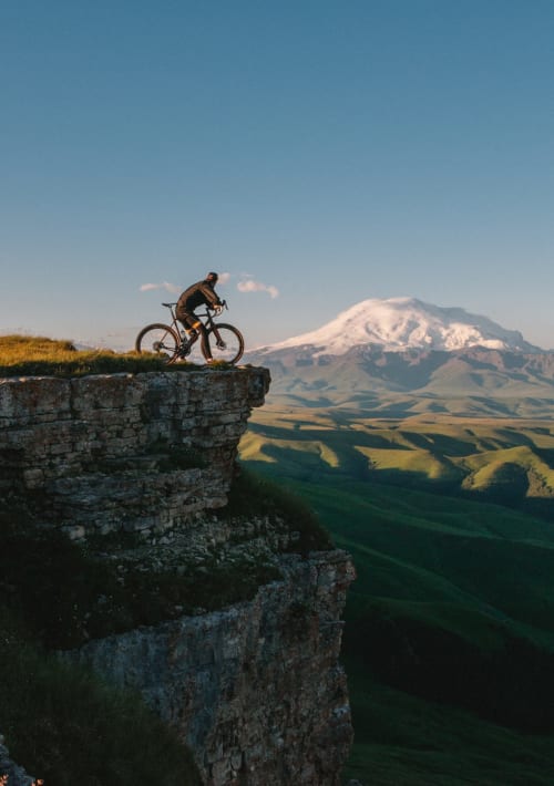 Mountainbiker am Klippenrand mit Blick auf schneebedeckten Vulkan und Tal bei Arvier