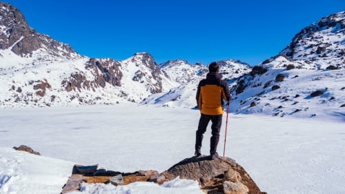 Wanderer mit orangenem Rucksack auf einem schneebedeckten Alpensee in der Nähe des Hotels Le Clou in Arvier, italienische Berge
