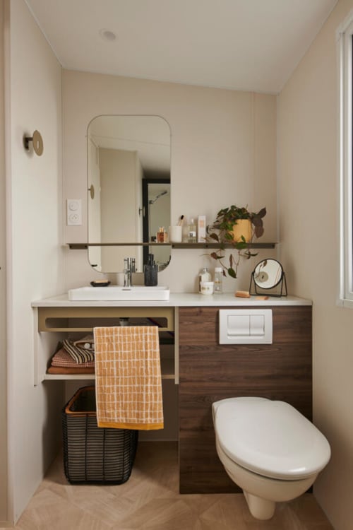 Salle de bain moderne de l'hôtel Le Maquis Plage avec meuble-lavabo en bois, miroir rond, plante en pot et toilettes blanches