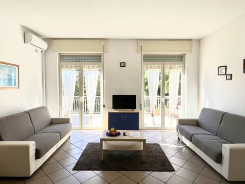 Luminoso soggiorno a Tirrenia con due divani grigi, TV, porte-finestre sul balcone e climatizzatore