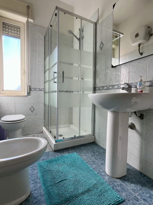 Bagno moderno dell'Apartment Tirreno con doccia in vetro, lavabo su colonna e tappetino turchese a Tirrenia