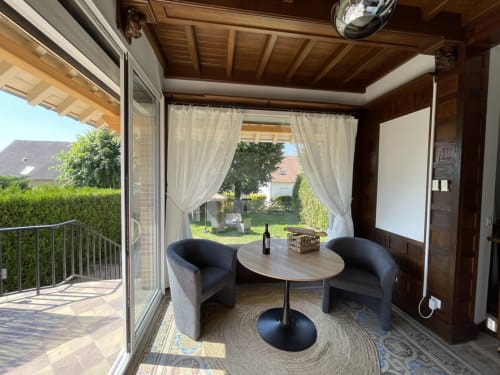 Véranda charmante avec vue sur le jardin, fauteuils noirs courbes, poutres en bois et vue sur les jardins luxuriants de cette maison d'hôtes française