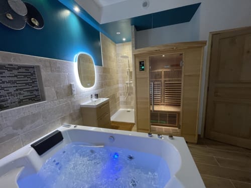 Salle de bain spa luxe avec miroir éclairé, jacuzzi blanc aux lumières bleues, sauna en bois et douche carrelée à Chartrettes