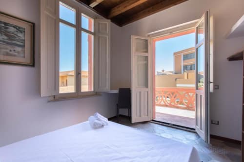 Camera da letto luminosa con biancheria bianca, travi in legno, finestra e porta che si aprono al balcone con vista sulla piscina a Maison Belle