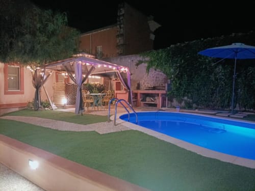 Piscina notturna del giardino di Maison Belle Époque con pergolato illuminato, ombrellone blu e area lounge a Oristano, Italia