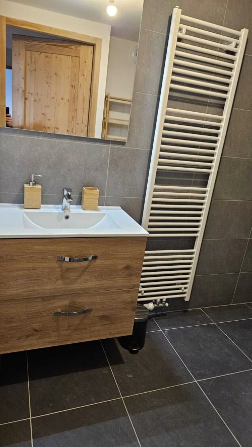 Salle de bain moderne à L'écrin des sapins : meuble vasque en bois, lavabo blanc, échelle porte-serviettes et carrelage foncé à Aime-la-Plagne.