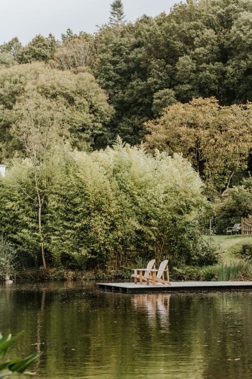 Wooden dock with Adirondack chairs on pond at Gîtes du Bulz in Pleyber-Christ, surrounded by lush trees