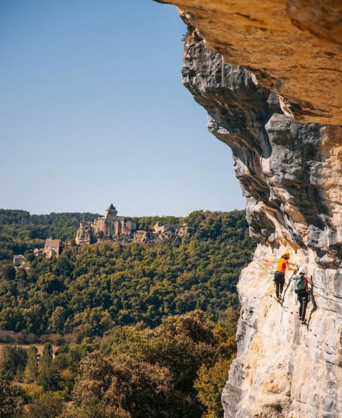 Grimpeur escaladant une falaise avec le château de Beynac visible dans la vallée boisée de la Dordogne, France