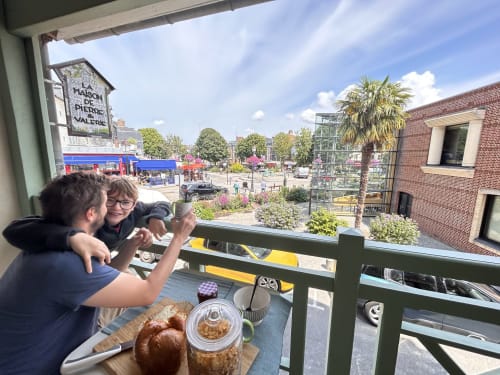 Couple enjoying breakfast on balcony overlooking Honfleur city square with pastries and coffee at Pierre et Valérie cottage.
