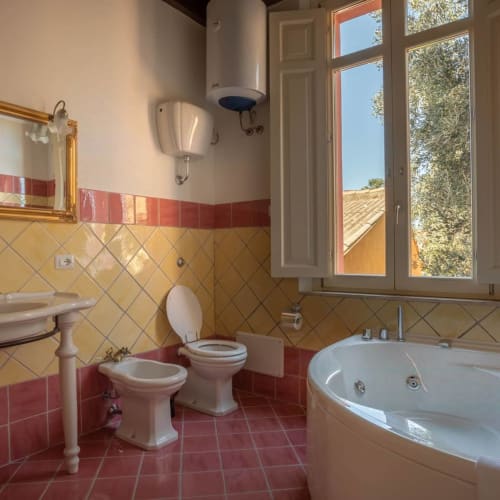 Bagno spazioso nell'appartamento B&B di Oristano con piastrelle rosse e gialle, bidet, toilet, vasca jacuzzi rotonda e vista sul giardino