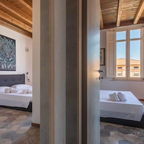 Camera da letto in appartamento Belle Époque con travi in legno a vista, pareti bianche e ampie finestre con vista su Oristano