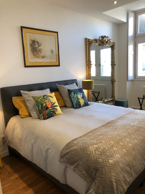 Chambre d'appartement à Beaune avec lit noir, coussins jaunes et bleu-canard, art mural lion encadré, miroir doré ouvragé et vue sur la ville