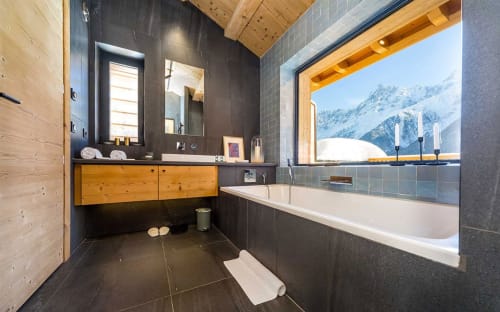 Salle de bain alpine moderne au Chalet Mont-Blanc avec vue sur les montagnes enneigées, murs en carrelage sombre, meuble vasque en bois et baignoire profonde