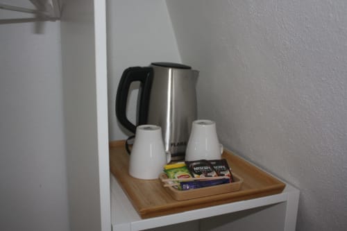 Estação de café e chá no quarto do hotel VAGO Alcântara Suites com chaleira elétrica, xícaras e sachês em bandeja de madeira