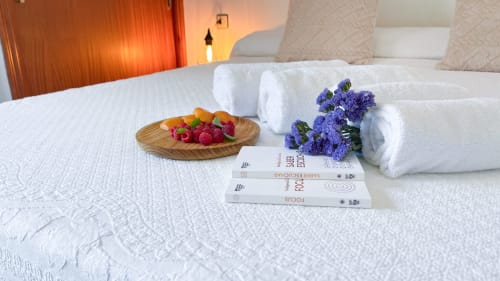 Lujosa habitación de villa con ropa de cama blanca, toallas enrolladas, flores moradas, frutas frescas y libros en Casa Rural Villa del