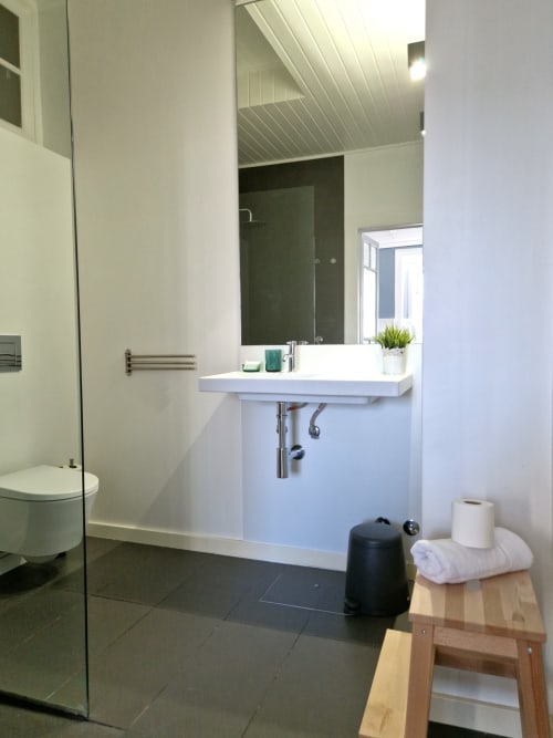 Banheiro moderno na VAGO Alcântara Suites Lisboa com lavatório branco, espelho escuro, box de vidro e banqueta de madeira com