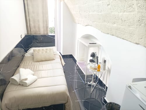Compact hotel room at Newhouse Bari with white bed, dark marble floor, small desk, fan, and white brick wall balcony view.