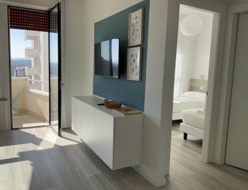 Modern apartment living room with teal accent wall, wall-mounted TV, white cabinetry, and balcony overlooking Misano