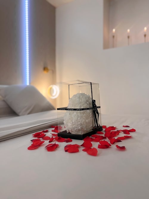 Un cœur blanc en pétales dans une boite cadeau, entourée de pétales de roses rouges éparpillées sur un lit.