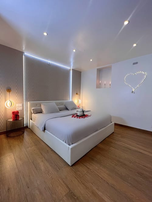 Chambre décorée pour des amoureux. Un cœur led au mur et un trio de bougeoir dans un renfoncement. Des pétales sur le lit King Size. 