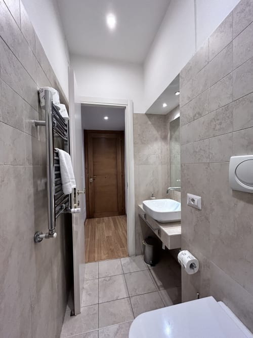 Modern bathroom at Maison Vaticana Rome with gray tile walls, heated towel rack, white sink, toilet, and wooden flooring.