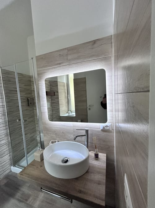 Modern bathroom at Maison Vaticana Rome with round white sink, LED mirror, wood-look tiles, and glass shower enclosure