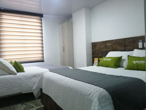 Habitación Hotel Maderos Cali con camas gemelas modernas, almohadas verdes, cabecera de madera oscura y persianas de ventana a rayas
