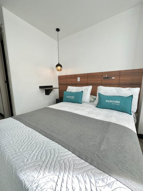 Habitación moderna en Hotel Maderos Cali con camas queen, almohadas Ayenda turquesas, cabecera de madera e iluminación colgante.