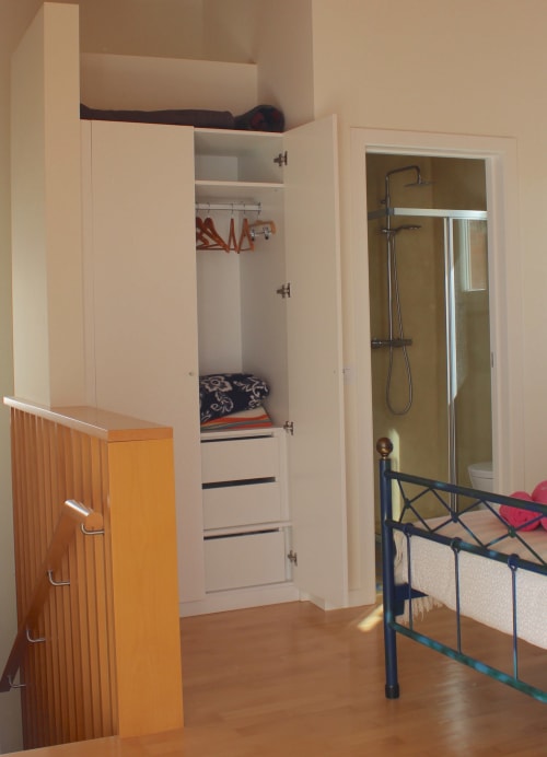 Quarto de estúdio com guarda-roupa branco, cama com estrutura metálica, móvel laranja e casa de banho privativa em Bordeira, Portugal.