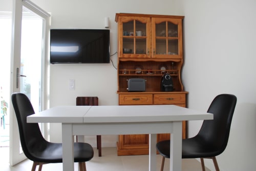 Área de jantar moderna no apartamento Estudio Hortas com mesa branca, cadeiras pretas, TV na parede e móvel em madeira