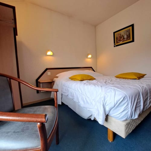 Chambre de l'hôtel Les Deux Porches à Verrières avec lit double, oreillers jaunes, chaise en bois et éclairage mural ambiant