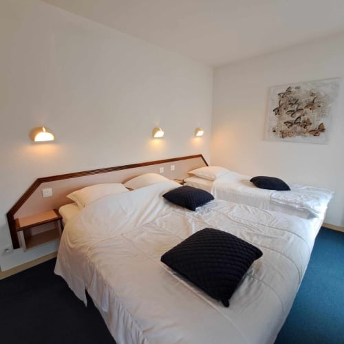 Chambre double de l'hôtel Les Deux Porches à Verrières avec literie blanche, oreillers noirs et éclairage mural chaleureux