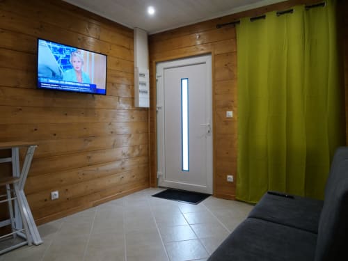Entrée de l'appartement Sci Saviya à Égletons : murs en bois, télévision écran plat, rideaux verts et canapé noir