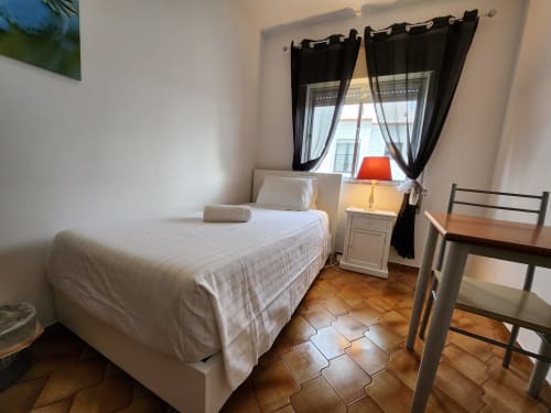 Quarto aconchegante com cama branca, piso de madeira e janela com cortinas pretas na Casa do Sol, Lagos, Portugal