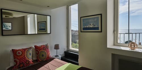 Chambre lumineuse à La Luna B&B Marseille avec coussins rouges à motifs, vue sur mer et œuvres d'art marseillaises sur murs olive