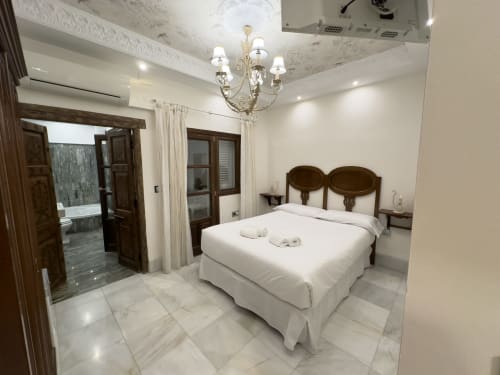 Dormitorio de lujo en Suite Comares Granada con suelos de mármol, araña elegante, muebles de madera y ropa de cama blanca