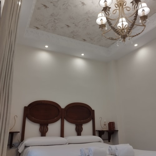 Habitación elegante en Suite Comares Granada con cabecero de madera oscura, lámpara de araña ornamentada y paredes blancas