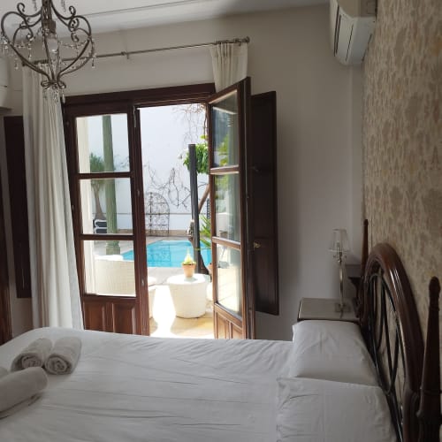 Dormitorio Suite Comares Granada con vista a piscina a través de puertas francesas, ropa de cama blanca y araña de hierro forjado