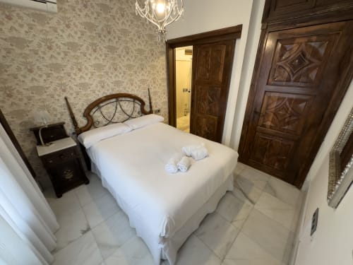 Elegante dormitorio en el hotel Suite Comares de Granada con cama de hierro forjado, papel tapiz floral y puertas de madera